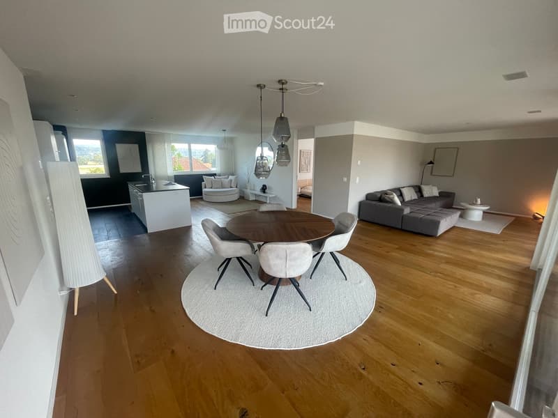 Open-plan living and dining area with wooden flooring, a round dining table with chairs, a grey sectional sofa, a small round seating area, and a modern kitchen counter.