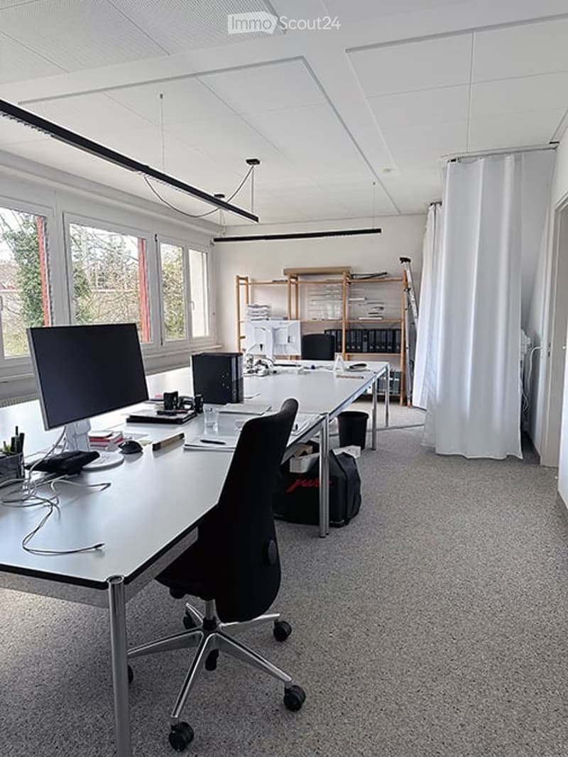 Modern office space with large windows, white walls, and a grey carpeted floor. A white desk holds a computer monitor, keyboard, mouse, and office supplies. An office chair is positioned in front of the desk. A black curtain is on the right side, and shel