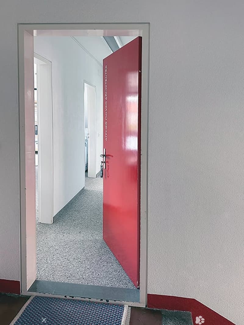 A clean, modern hallway with a red door at the end, light grey walls, and a grey carpeted floor.