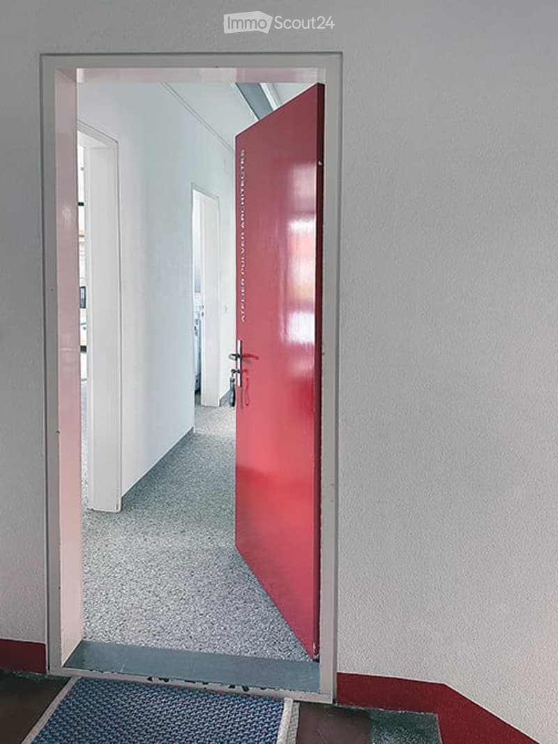 A clean, modern hallway with a red door at the end, light grey walls, and a grey carpeted floor.