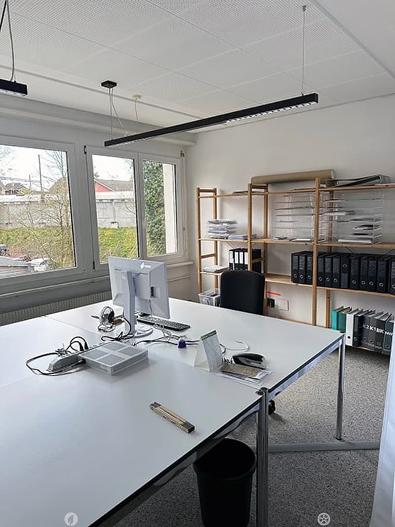 Office space with a large desk, computer setup, chair, shelves with books and files, and a window view of the outside.