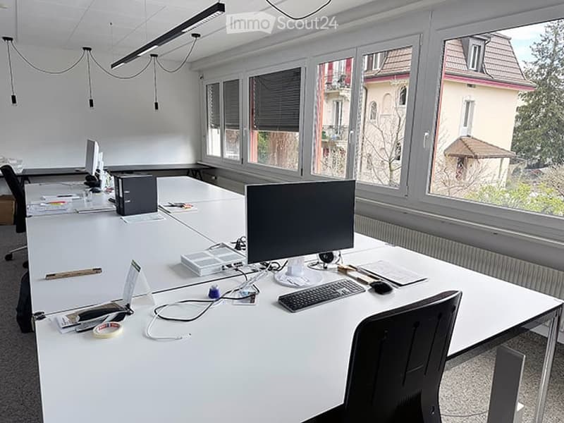Bright office space with large windows, white desks, chairs, and computer equipment.