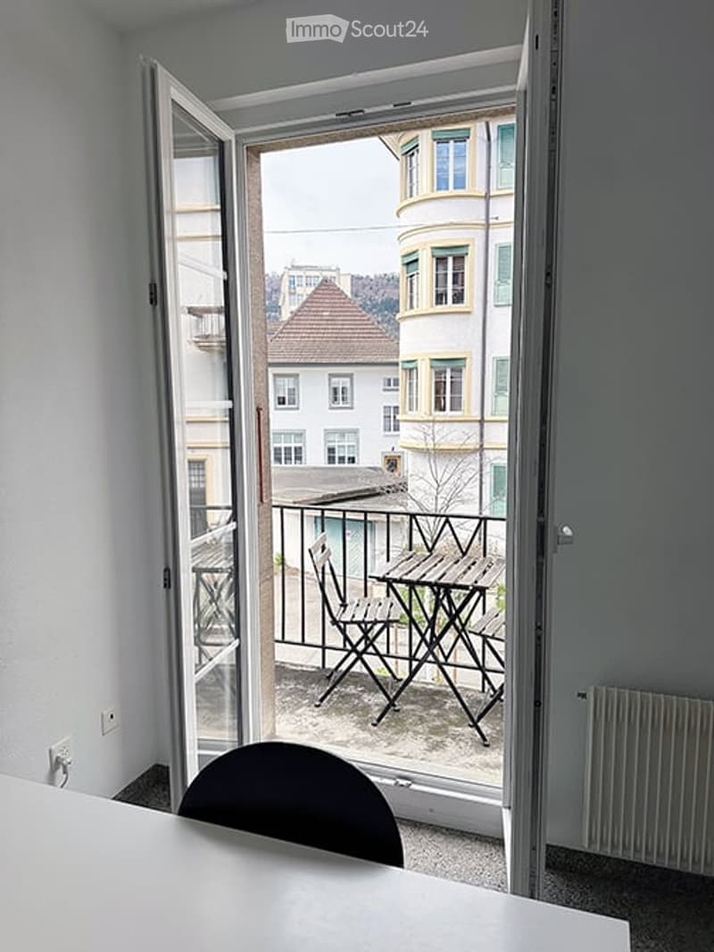 Open balcony door leads to a small outdoor seating area with a table and chairs, overlooking a cityscape with buildings and a sloped roof.
