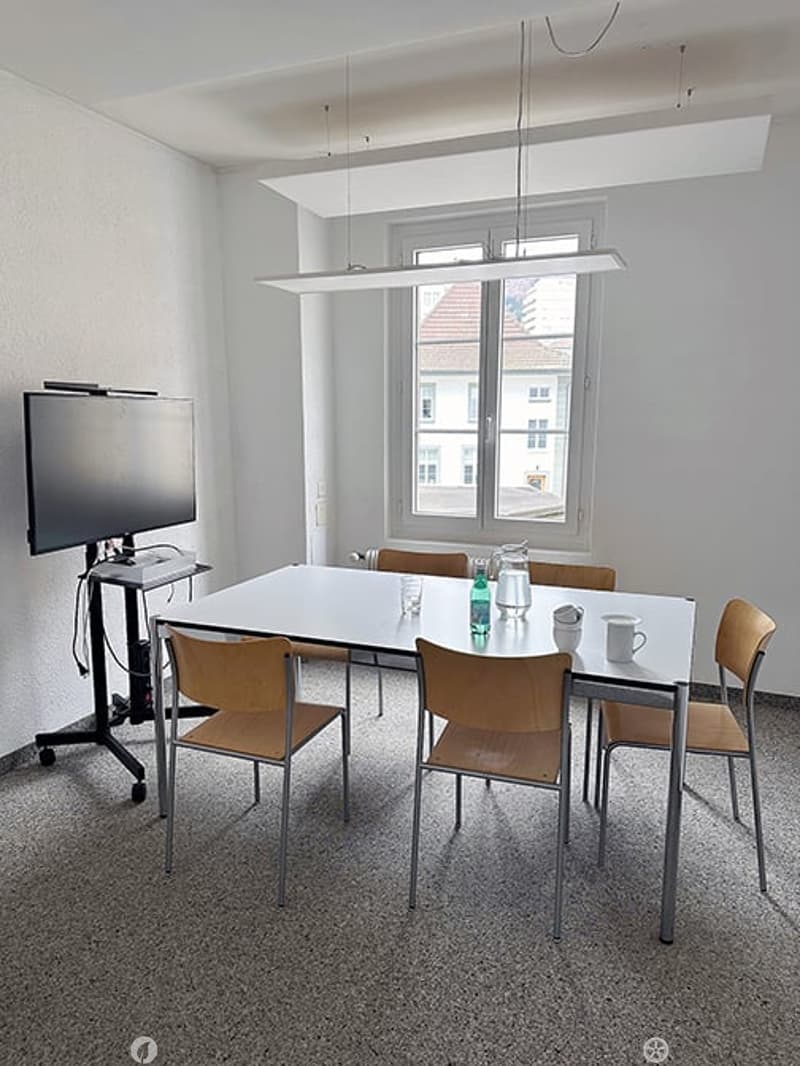 Bright room with a large white table surrounded by six chairs, a screen on a stand, and a window with a view of a building outside.