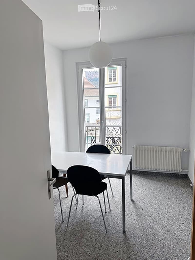 A small, unfurnished room with a white table and two black chairs, large windows with a view of the outside, and a radiator on the wall.