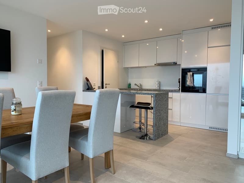 A modern, open-plan kitchen and dining area with white cabinets, a wooden dining table, grey chairs, and a granite countertop. The kitchen features a stove, sink, and built-in oven, with a small bar stool at the counter.