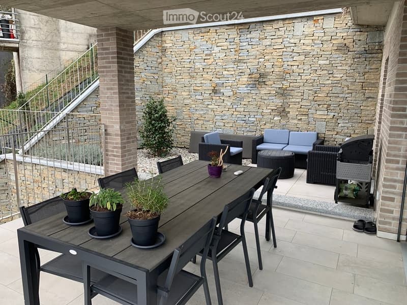 Modern outdoor patio with a stone wall backdrop, featuring a dining table with chairs, potted plants, and a seating area with wicker chairs and a sofa.