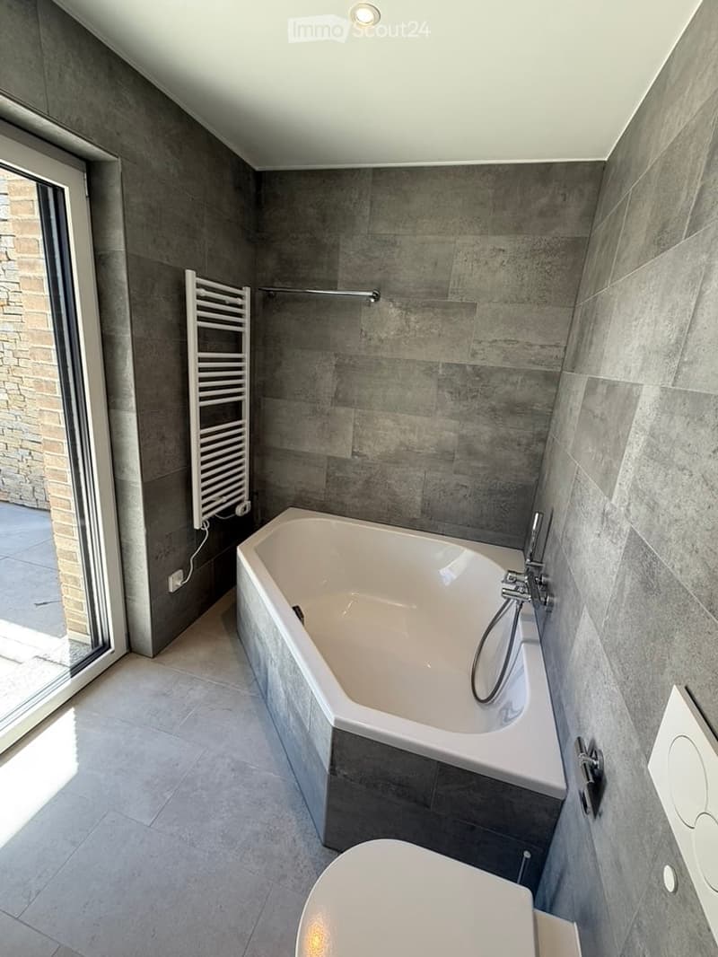Modern bathroom with a large bathtub, toilet, radiator, and sleek grey tiles.
