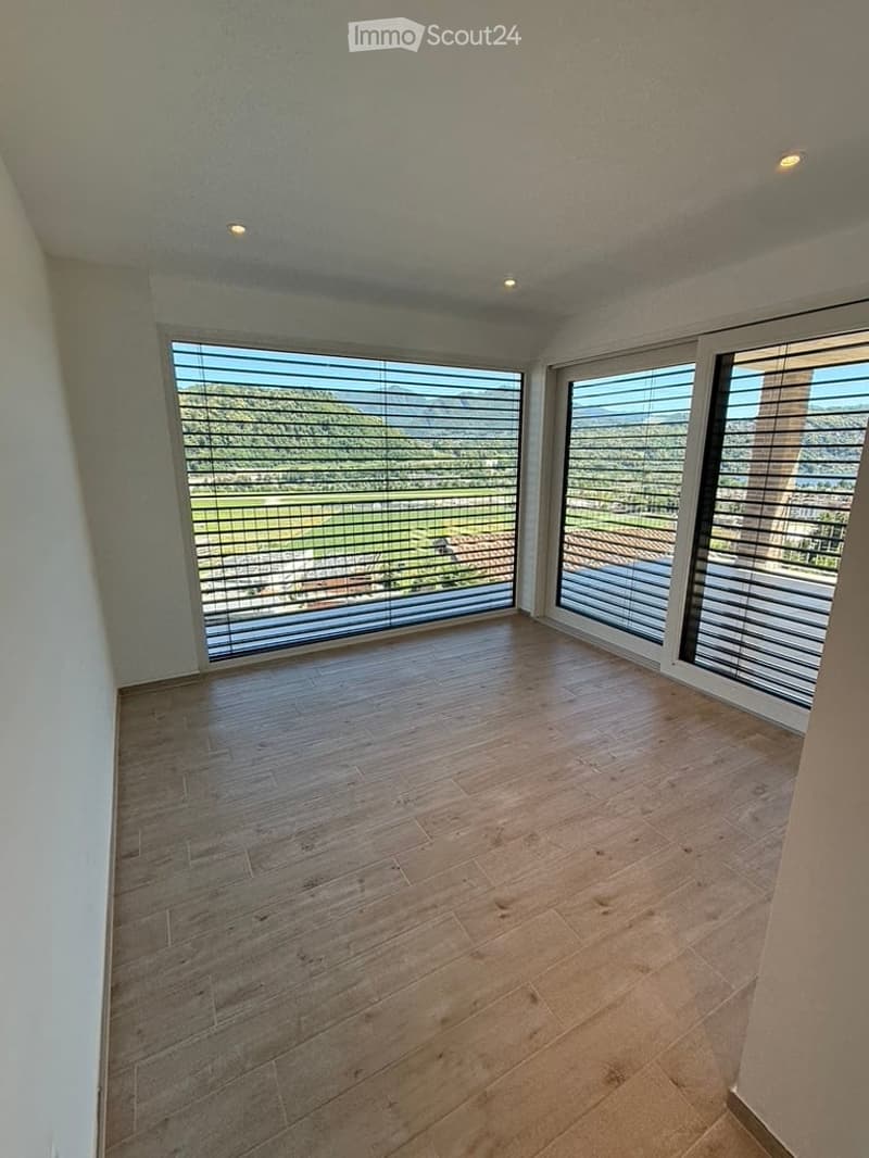 Empty room with wooden flooring, large windows with blinds, and a scenic view of the landscape.