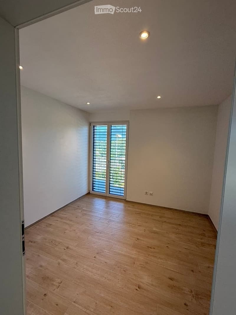 Empty room with wooden flooring, white walls, and a large window with blinds. The room is well-lit with recessed ceiling lights.