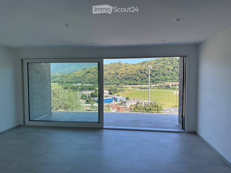 Empty room with large sliding glass doors opening to a balcony, offering a scenic view of a green landscape and hills.