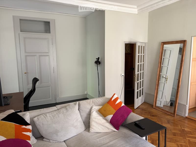Living room with a grey sofa, colorful pillows, a small black side table with a remote control, a wooden-framed mirror, and a black office chair near the door.