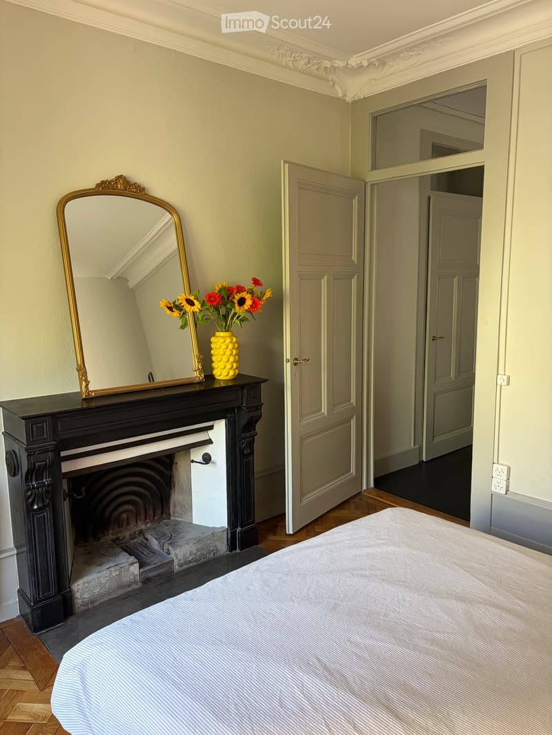 Elegant bedroom with a large bed, ornate fireplace, decorative mirror, and vase with flowers on the mantel.