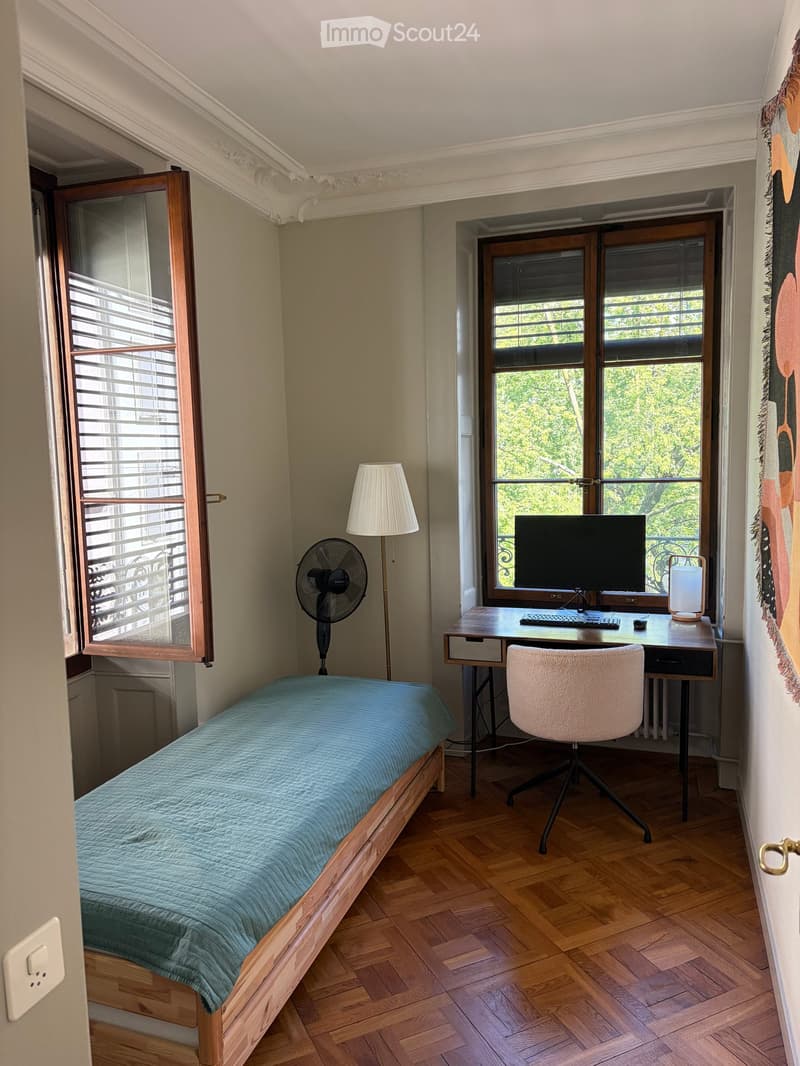 A small, well-lit room with a single bed, a desk with a computer, a chair, a fan, and a lamp. The room has wooden flooring and large windows with blinds.