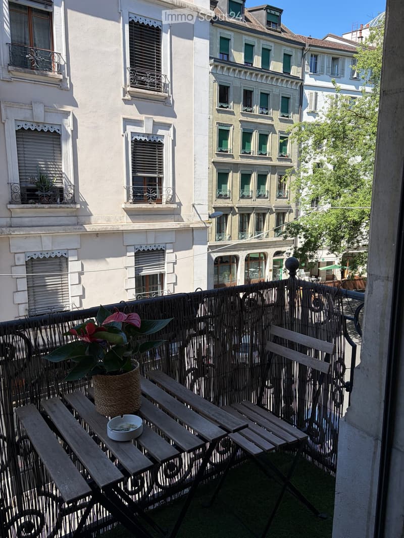 Balcony with wooden table, chairs, and a potted plant, overlooking a city street with historic buildings.
