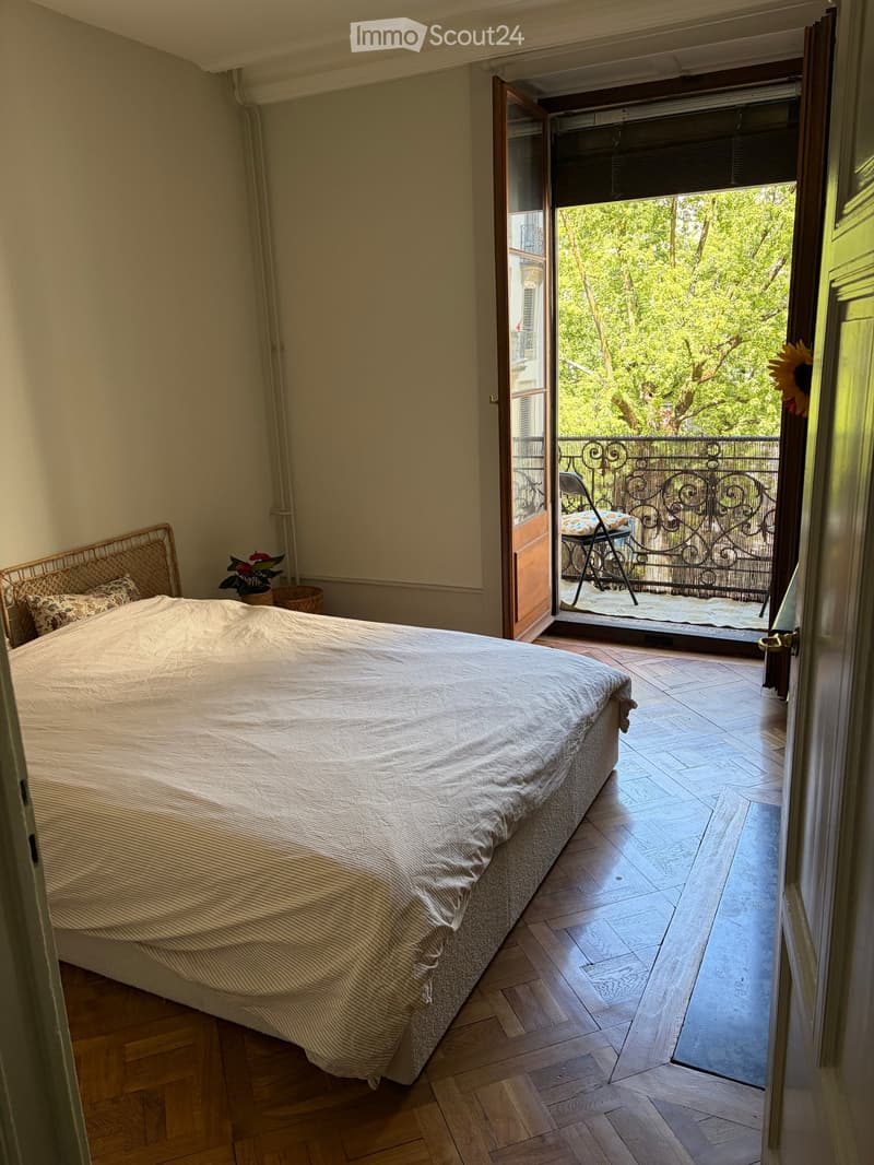 Cozy bedroom with wooden flooring, a large bed, a small balcony with a view of greenery, and a small table and chair on the balcony.