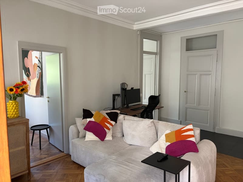 A modern living room with a white sofa, colorful pillows, a small table, a computer setup, and a vase with sunflowers on a side table.