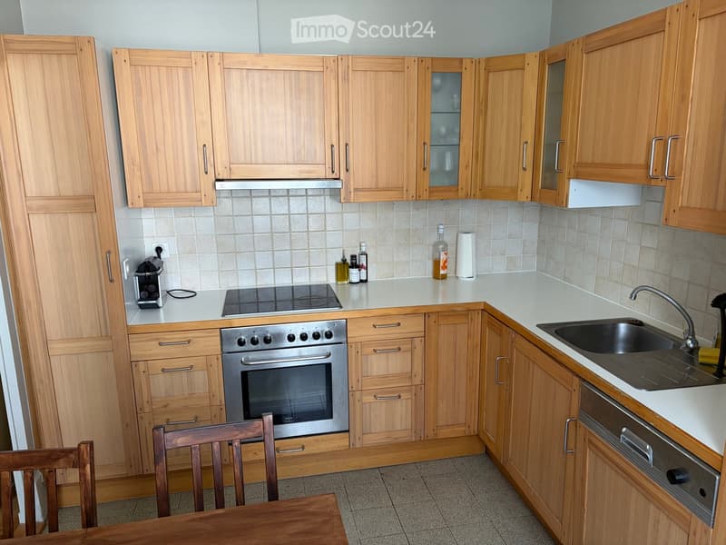 Modern kitchen with light wooden cabinets, stainless steel oven, sink, and countertop items including a coffee maker and bottles.