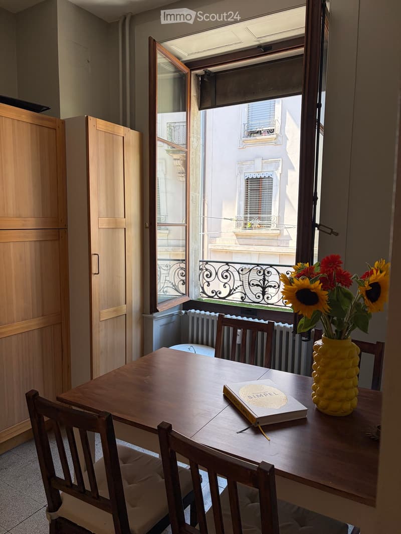 A small, cozy room with a wooden table, two chairs, a vase with sunflowers, and a book on the table. The room has large windows opening to a balcony with a view of the street.