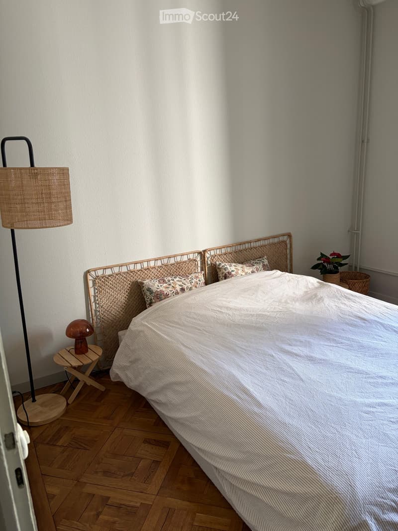 Cozy bedroom with a wicker bed frame, striped bedding, wooden floor, floor lamp, small wooden stool with table lamp, and a potted plant near the window.