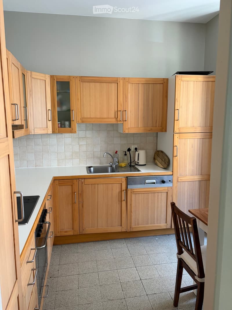 A compact kitchen with light wooden cabinets, a sink, dishwasher, oven, and a small dining area with a chair and table.