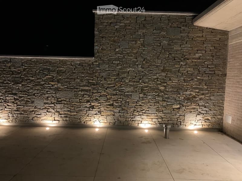 An outdoor patio area with a stone wall backdrop and integrated lighting, featuring a clean tiled floor and a small metallic trash can.