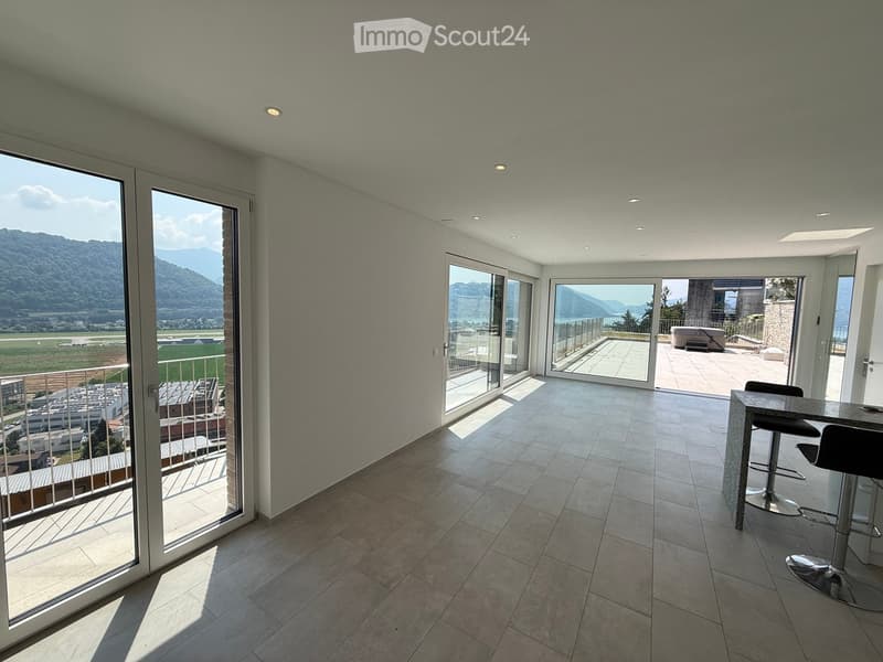 Modern, open-plan room with large glass doors leading to a balcony, tiled floor, bar counter with two stools, and scenic mountain view.