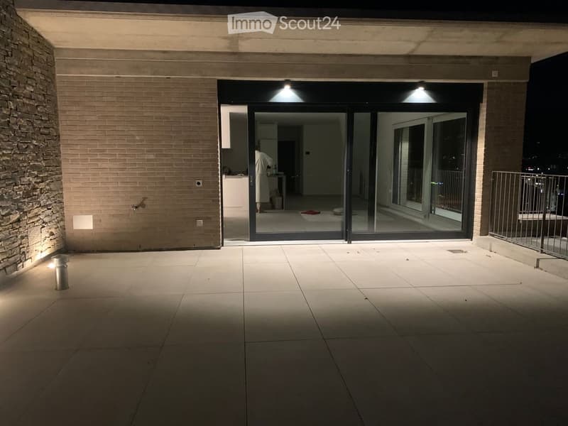 Nighttime view of a modern balcony with large glass sliding doors leading to an interior space, tiled floor, and ambient lighting.