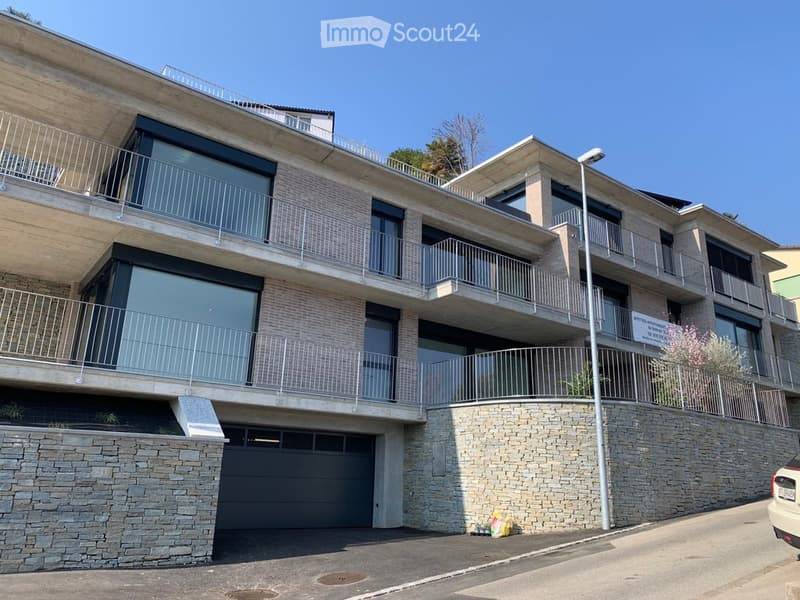 Modern multi-story building with balconies, large windows, and a stone facade. Features include a garage and a raised ground floor.