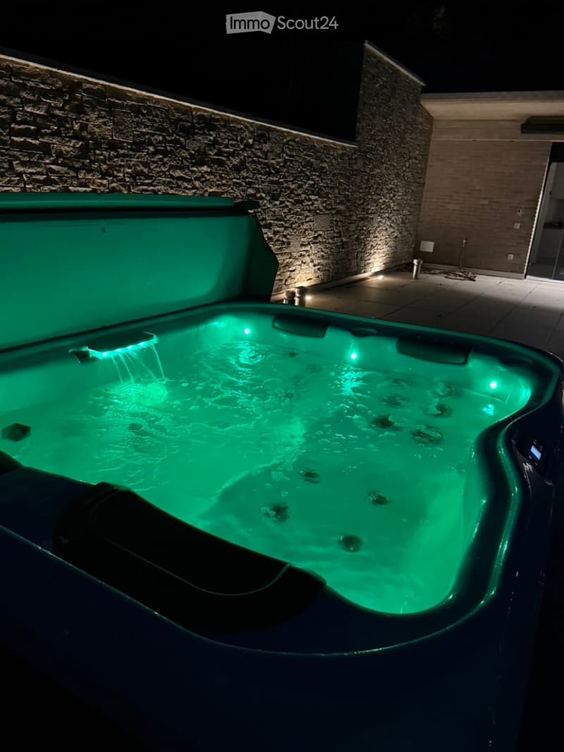 Nighttime view of a modern outdoor hot tub with green lighting, surrounded by a stone wall and illuminated pathway.