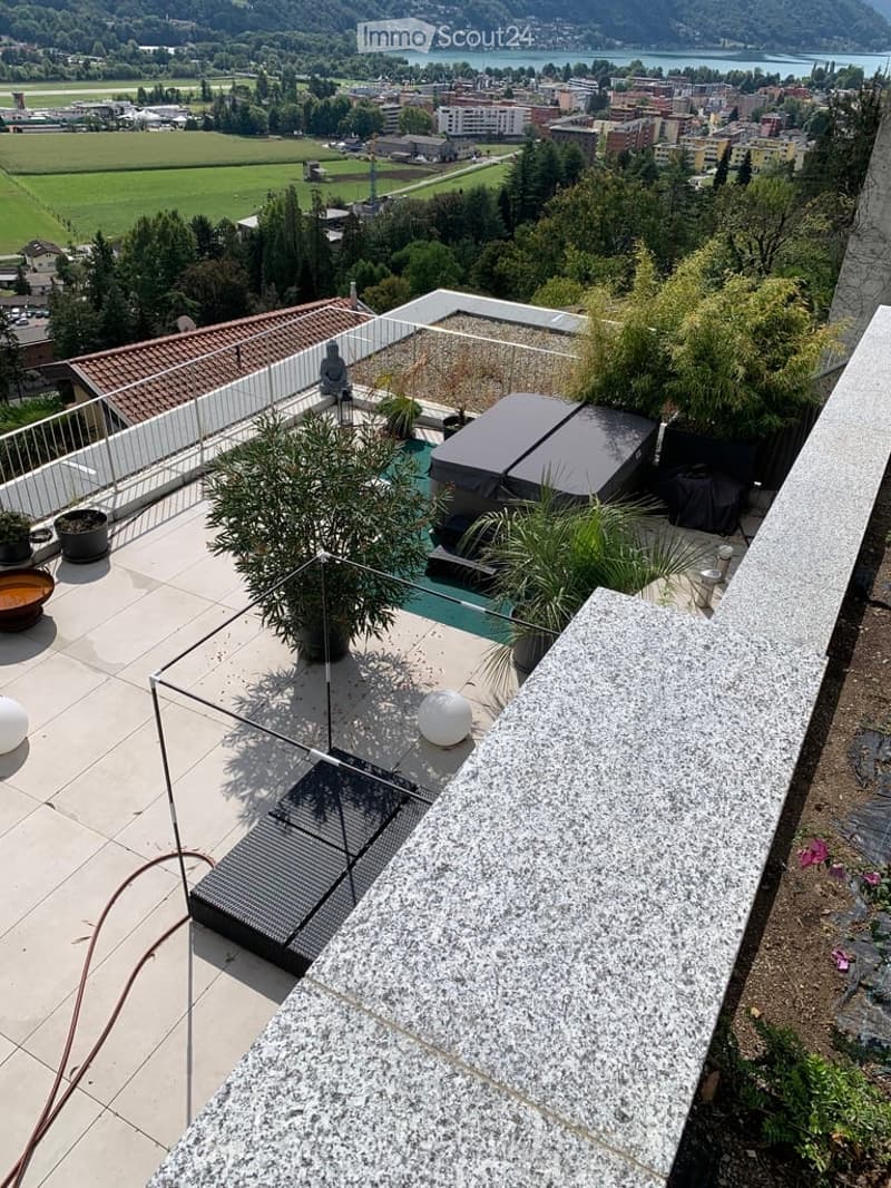 Rooftop terrace with modern glass railing, lounge chairs, potted plants, and scenic view of a lake and town.