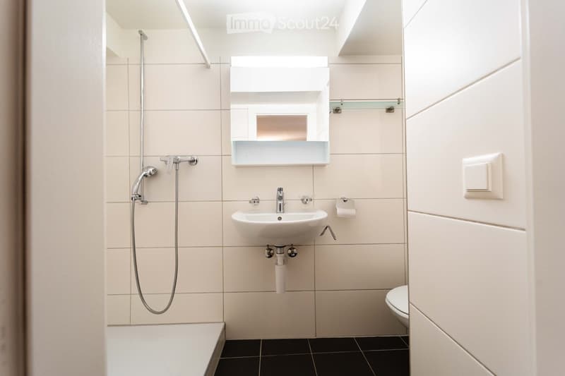 Modern bathroom with a white sink, mirror, toilet, and shower head. The walls are tiled in light beige, and the floor is black.