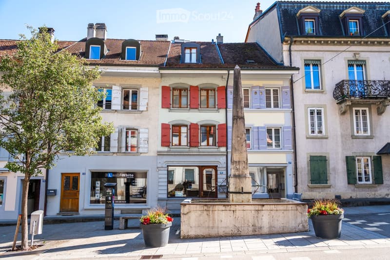 A row of historic buildings with varied facades, including red shutters, white shutters, and green shutters, with a central obelisk and surrounding planters.