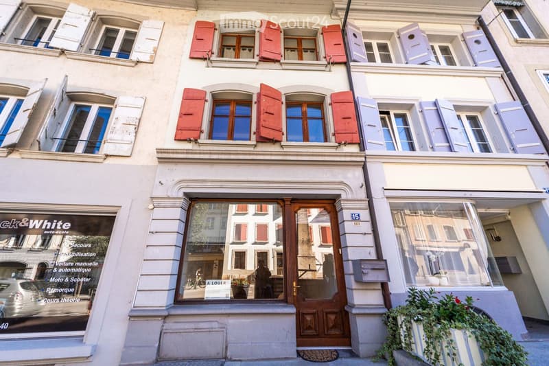 A multi-story building with classic European architecture, featuring red and blue shutters, large windows, and a storefront with glass windows and a wooden door.