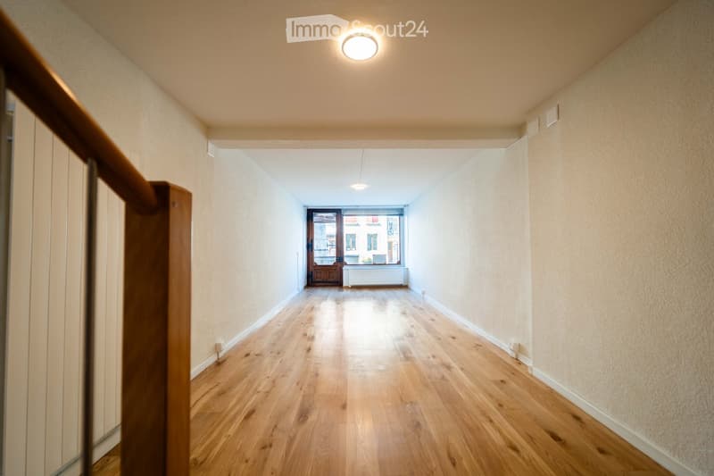 A well-lit hallway with wooden flooring, light-colored walls, and a staircase on the left. A door at the end leads to a room with a window and a small bench.