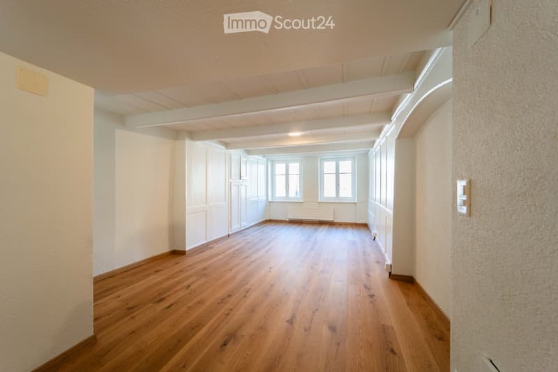 Empty hallway with wooden flooring, white walls, and large windows at the end.