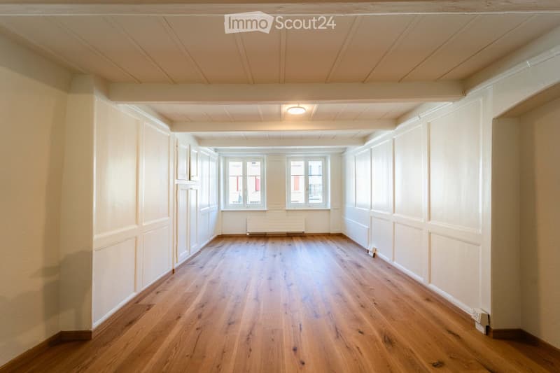 Empty hallway with wooden flooring, white walls, and large windows at the end. No furniture or equipment present.