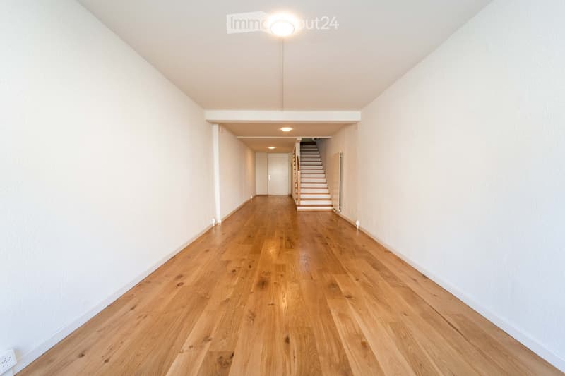 Empty hallway with light wooden flooring, white walls, and recessed lighting. A staircase leads upwards in the background.