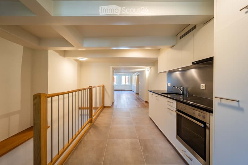 Modern hallway with wooden flooring, white walls, and a small kitchen area visible on the right side. The hallway features a wooden railing and a skylight above.