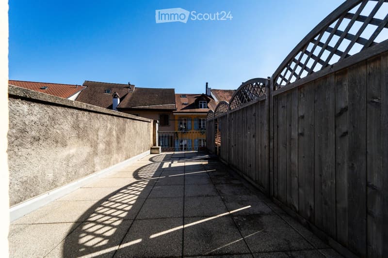 A narrow, paved pathway bordered by a tall concrete wall on one side and a wooden gate on the other, leading to a view of neighboring buildings with red-tiled roofs.