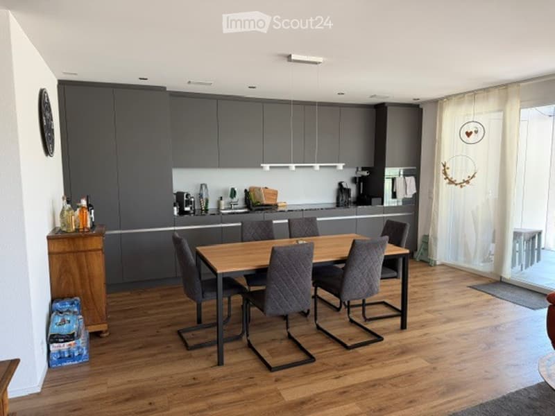Modern kitchen with grey cabinetry, a wooden dining table surrounded by grey chairs, and various kitchen appliances and items on the counter.