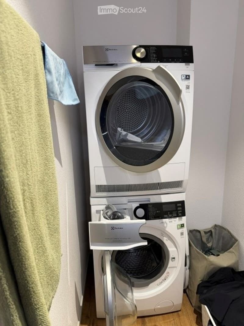 A compact bathroom with a stacked washing machine and dryer unit, a light blue towel hanging on the wall, and a small laundry basket on the floor.