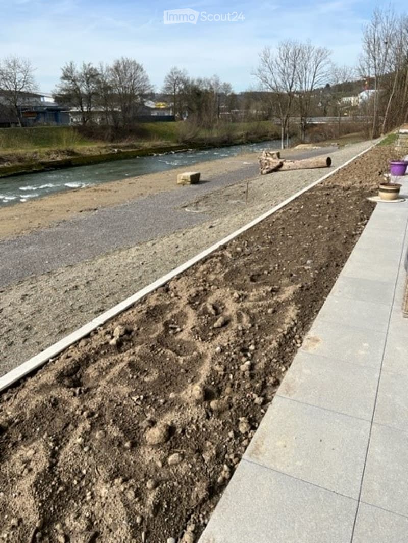 An outdoor area near a riverbank with gravel, soil, and a few scattered logs and bricks. The area is bordered by a paved walkway and has a view of the river and distant buildings.