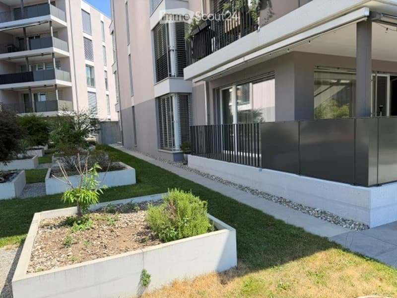 Modern residential complex with a small landscaped garden area featuring raised planters, low shrubs, and a neatly trimmed lawn.