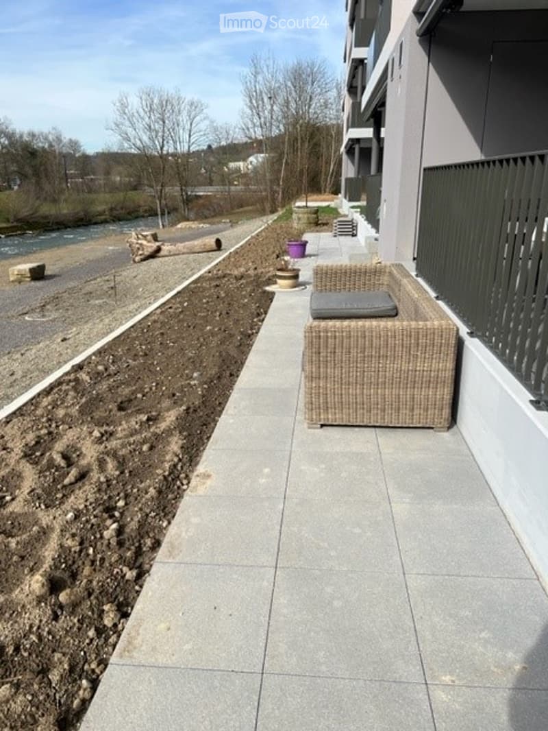 Balcony with a wicker chair and cushion, overlooking a landscaped area with a river and trees in the background.