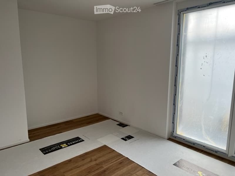 Empty room with wooden flooring and white walls, showing protective plastic sheets on the floor and a large window with frosted glass.
