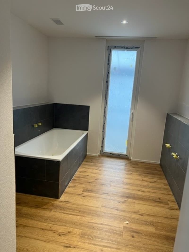 Modern bathroom with a freestanding bathtub, dark grey tiles, wooden flooring, and a frosted glass shower enclosure.