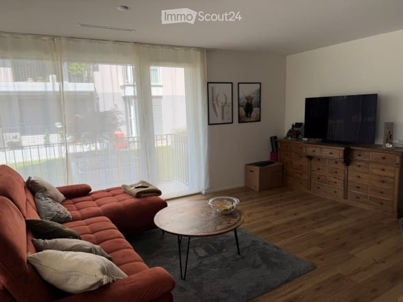 Modern living room with a red sectional sofa, wooden coffee table, flat-screen TV on a wooden dresser, and large windows with sheer curtains.