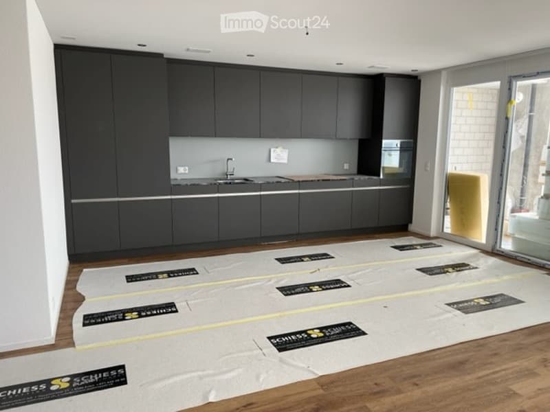 Modern kitchen with dark grey cabinets, a sink, and a wooden floor. Protective plastic sheets are laid out on the floor, likely for renovation or installation purposes.