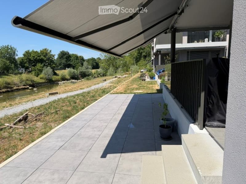 Balcony with a retractable awning, tiled floor, small potted plant, and a view of a river and greenery.